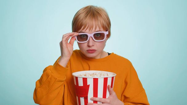 Jeune femme excitée dans des lunettes 3D manger du pop-corn et regarder la série télévisée intéressante, jeu de sport, film, contenu de film de médias sociaux en ligne. Blonde fille aux cheveux courts isolé sur fond de studio bleu - Photo, image