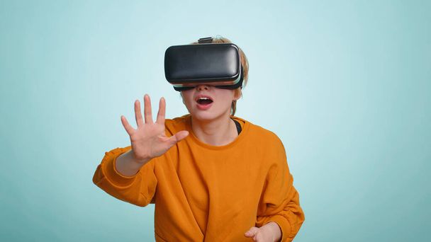 Γυναίκα που χρησιμοποιεί φουτουριστική τεχνολογία εικονικής πραγματικότητας κράνος ακουστικών VR app για να παίξει προσομοίωση 3D 360 video game, βλέποντας ταινία. Ξανθιά κοντή κοπέλα απομονωμένη σε μπλε φόντο στούντιο, σε εσωτερικούς χώρους - Φωτογραφία, εικόνα