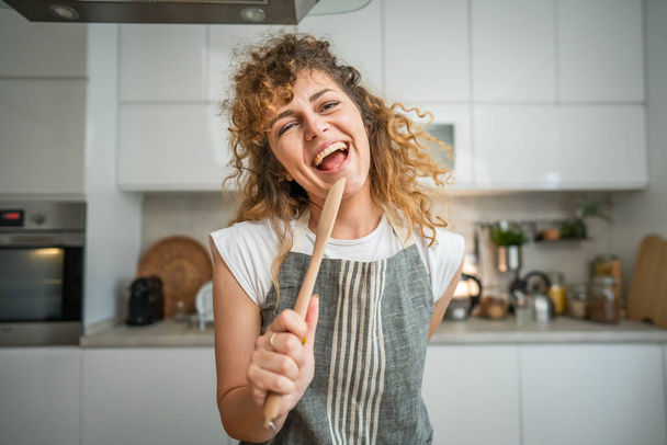 One happy young adult caucasian woman wear apron in the kitchen smile hold mixing spoon sing - Foto, immagini