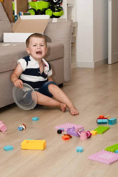 A small Caucasian beautiful boy was upset and crying while sitting on the floor with scattered toys. He does not want to remove the childrens multi-colored constructor. Concept - Photo, Image
