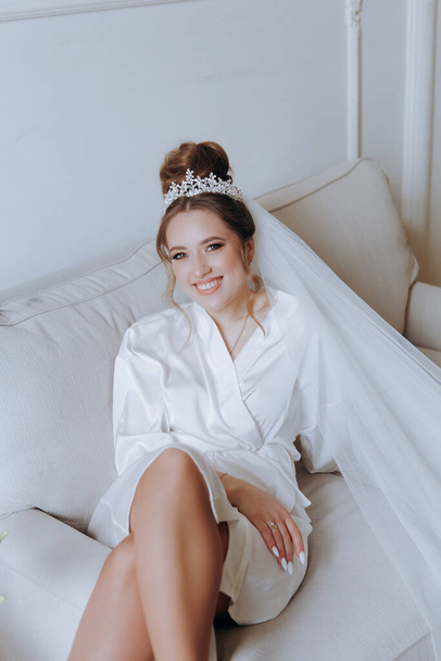 Morning preparations of a beautiful, sexy bride in a bathrobe on the balcony and on the sofa, where she puts on shoes in a wedding dress - Фото, изображение