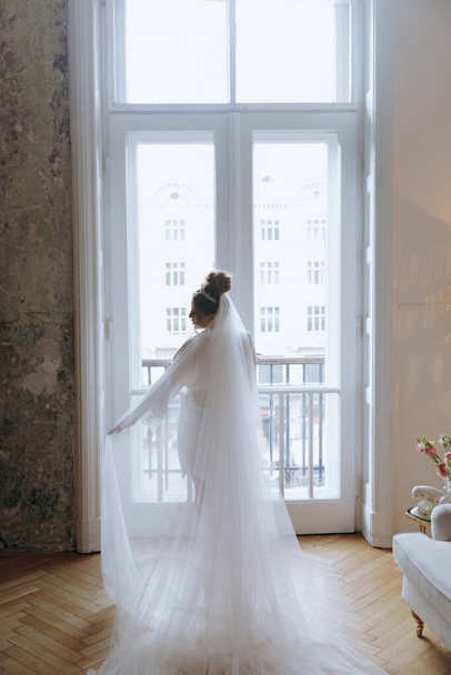 Morning preparations of a beautiful, sexy bride in a bathrobe on the balcony and on the sofa, where she puts on shoes in a wedding dress - Foto, Bild