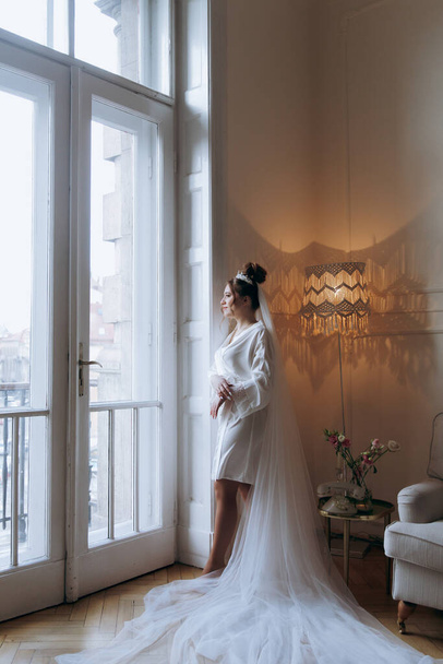 Morning preparations of a beautiful, sexy bride in a bathrobe on the balcony and on the sofa, where she puts on shoes in a wedding dress - Foto, immagini