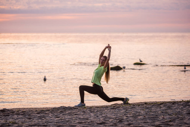 Young woman in sportswear does sit-ups early in morning on seashore at sunrise. Girl of slim build with long hair is stretching. Outdoor sports by the water. Fitness classes, body care, mental health - Foto, Bild