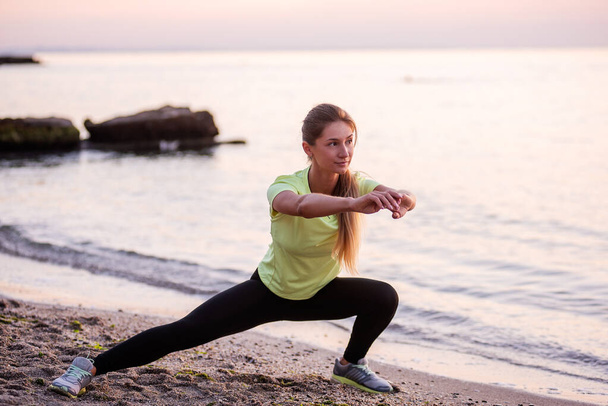 Young woman in sportswear does sit-ups early in morning on seashore at sunrise. Girl of slim build with long hair is stretching. Outdoor sports by the water. Fitness classes, body care, mental health - Photo, Image