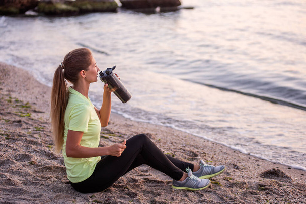 Young woman in sportswear is sitting on the seashore at sunrise. Girl is resting after workout, replenishes her water balance, drinks from a sports bottle, looks at the water. Morning outdoor fitness classes. - Foto, Imagen