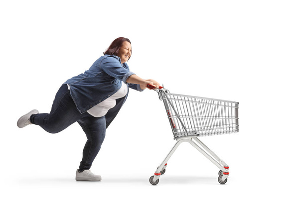 Full length profile shot of a corpulent young woman running with an empty shopping cart isolated on white background - 写真・画像