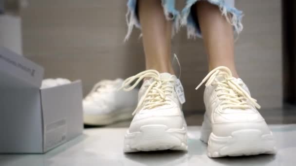 Mädchen mit Nahaufnahme ihrer Beine und probiert Schuhe in einer Modeboutique an. Sie wählt weiße Turnschuhe, um viele Schachteln und Regale mit Styles herum. Schuhe kaufen und anprobieren. - Filmmaterial, Video