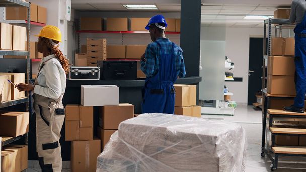Αφροαμερικάνος επόπτης κοιτάζει τα αγαθά σε πακέτα, δουλεύοντας με διάφορους υπαλλήλους για να στείλει εμπορεύματα. Νεαρή γυναίκα διευθυντής σε φόρμες ελέγχου προϊόντων παραγγελίας για logistics. - Φωτογραφία, εικόνα