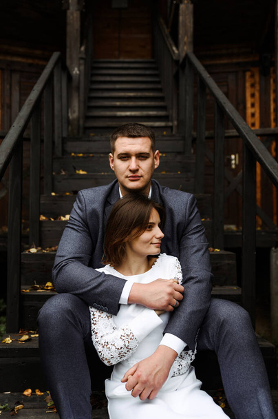 young wedding couple sits on a wooden staircase against the backdrop of an ecological house in the autumn season. newlyweds on the background of a wooden house in rustic style. - Fotó, kép