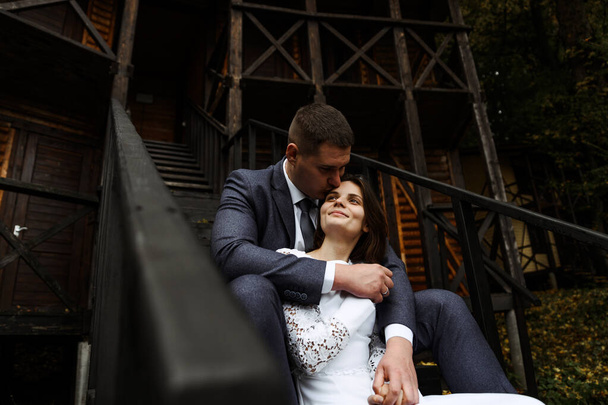 young wedding couple sits on a wooden staircase against the backdrop of an ecological house in the autumn season. newlyweds on the background of a wooden house in rustic style. - 写真・画像
