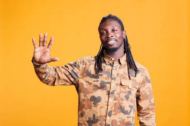 Joyful african american man greeting friend with wave in studio over yellow background, having positive facial expression. Cheerful young adult doing salute gesture to greet people. Hi or goodbye sign - Photo, Image