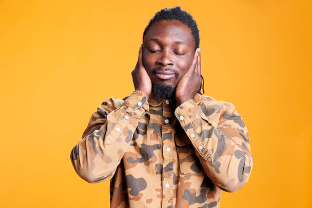 Man with close eyes covering ears with palms, doing three wise monkeys gesture in front of camera over yellow background. African american person not listening to noise and not speaking in studio - Photo, Image
