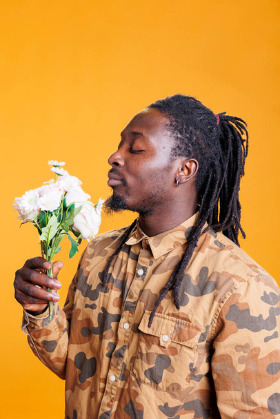 Portrait of joyful man enjoying white roses smell, posing with flowes for valentine s day in studio over yellow background. African american romantic young adult celebrating love holiday - Foto, Imagen