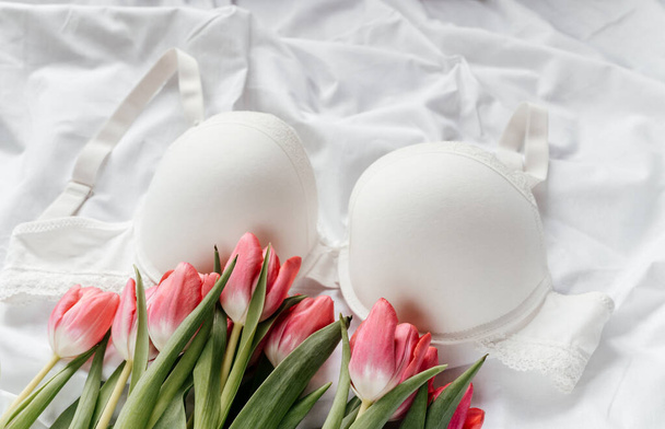 Gentle white lace bra on the bed with tulips. Women tender lingerie, underwear. Top view, close up. Flat lay, beauty blog or social media minimal concept. Present for Valentines, Womens day - Photo, Image