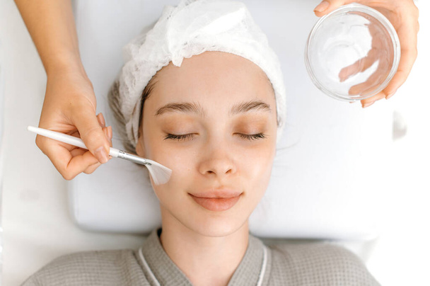 Top view. Facial skin care procedures in a beauty salon. The cosmetologist cleans the face of a beautiful caucasian woman with special gel before applying a mask, peeling, scrub - Photo, image