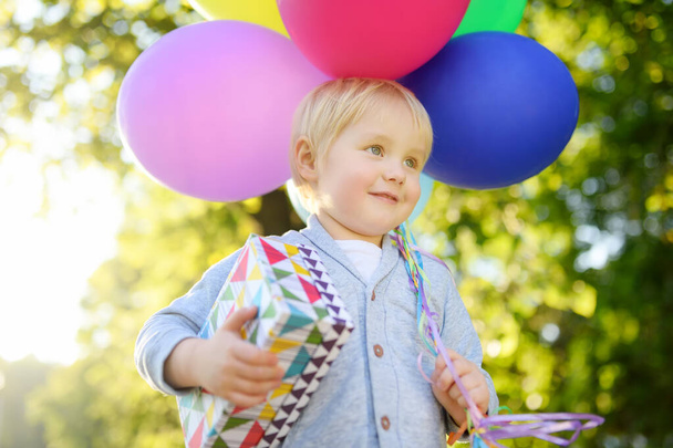 Cute little child going to congratulate a friend on his birthday. Toddler holding bundle of colorful balloons and gift in festive box. Preschoolers baby celebrating outdoor party in sunny summer park. - Foto, Imagen