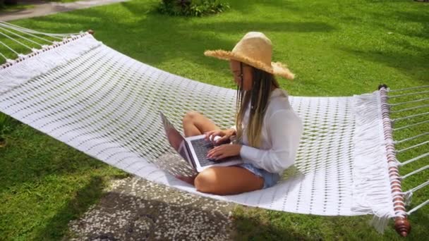Hasır şapkalı kadın programcı, yaz tatilinde hamakta otururken bilgisayarla çevrimiçi iş yapıyor. Kadın bilişim programcısı dijital göçebe seyahat ederken dizüstü bilgisayarını serbest çalışmak için kullanıyor. - Video, Çekim