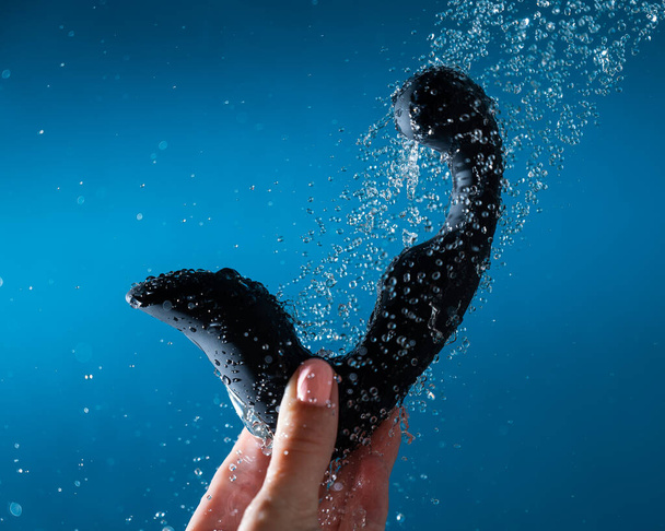 A woman washes a black prostate stimulator under running water on a blue background. Sex toy hygiene concept - 写真・画像
