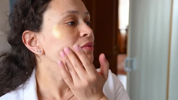 Close-up kobieta w średnim wieku z piegami, z hydrożel plastry pod oczami, stosując krem nawilżający i masujący twarz z nefrytowym kamiennym masażem rolki w łazience w domu. Antystarzeniowe zabiegi spa - Materiał filmowy, wideo