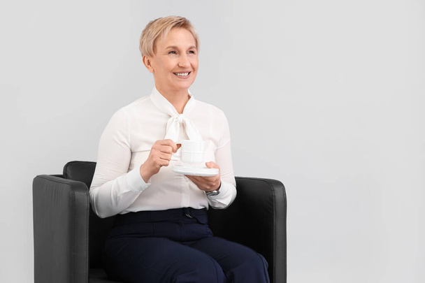 Madura mujer de negocios bebiendo café mientras se sienta en sillón sobre fondo gris - Foto, Imagen