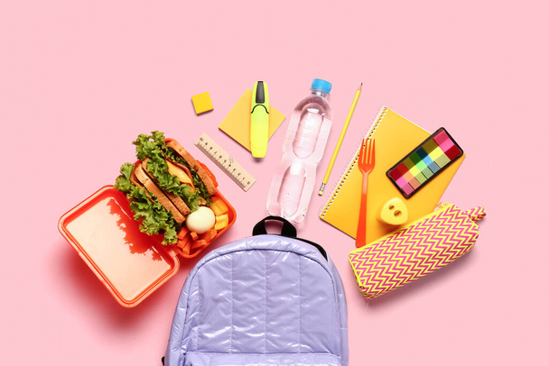 Plecak, papeteria, butelka wody i pudełko śniadaniowe ze smacznym jedzeniem na różowym tle - Zdjęcie, obraz