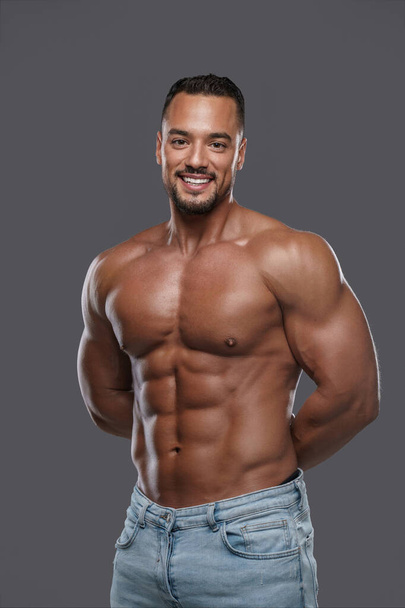 Charming and muscular male model with a dazzling smile poses shirtless against a neutral grey backdrop, emanating confidence and sex appeal - Foto, Imagen