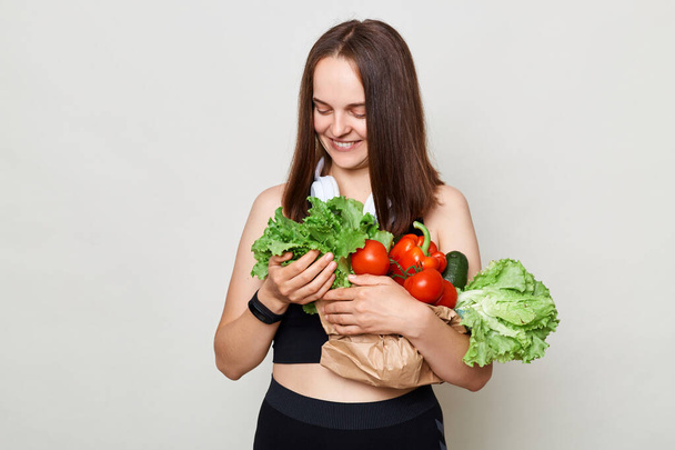 Cute calm smiling woman with dark hair carries vegetables bought at market isolated over gray background looking at green organic lettuce eating healthy food. - Foto, afbeelding