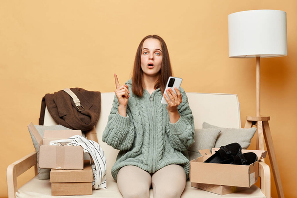 Εμπνευσμένη γυναίκα φορώντας πλεκτό πουλόβερ με παραγγελθέντα ρούχα σε κουτιά που κάθονται στο σπίτι στον καναπέ ενάντια σε μπεζ τοίχο χρησιμοποιώντας smartphone έθεσε δάχτυλο έχοντας ιδέα. - Φωτογραφία, εικόνα