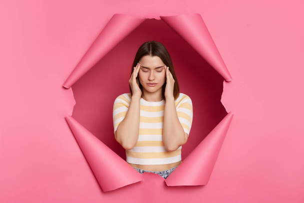 Sick unhealthy brunette woman wearing striped t shirt posing in torn pink paper wall suffering headache massaging temples grimacing frowning face. - Photo, image