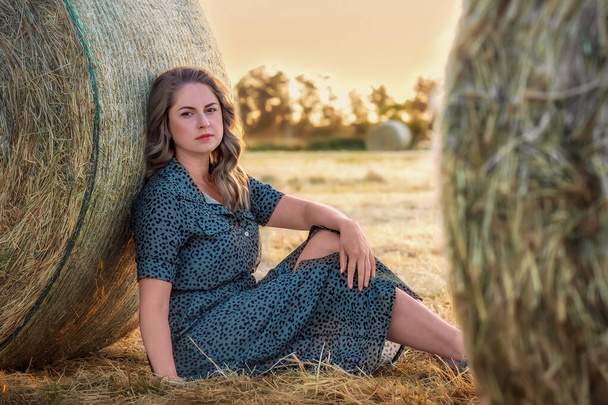 Красива молода жінка у літньому вбранні сидить у полі, спираючись на сіна. - Фото, зображення