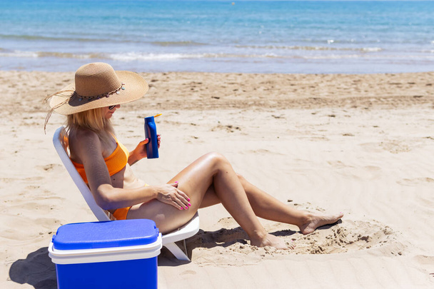 een gebruind meisje in een badpak zit op een ligstoel smeert zonnebrandcrème op haar benen zijaanzicht op het strand en er is een blauwe draagbare koelkast voor drankjes in de buurt. Zomervakantie Hoge kwaliteit foto - Foto, afbeelding