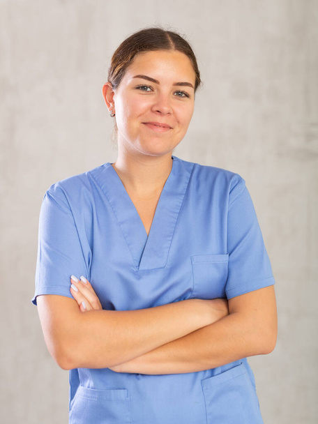 Friendly young nurse in blue coat posing against gray studio background, looking at camera with smile - Foto, Imagen