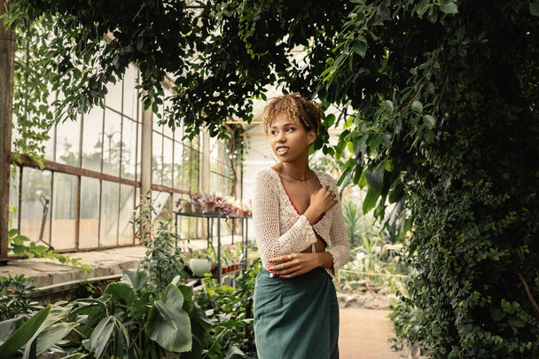 Mosolygós és divatos fiatal afro-amerikai nő nyáron kötött top és szoknya néz félre, miközben áll közelében zöld növények a kerti központban, elegáns nő élvezi buja trópusi környezetben - Fotó, kép