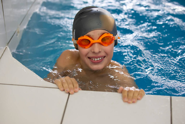 プールの中の水泳帽とゴーグルの少年。彼は笑顔でカメラを覗き込む。スポーツプールの子供. - 写真・画像
