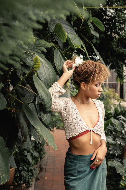 Mujer afroamericana joven de moda en la parte superior de punto y falda tocando el cabello y de pie cerca de follaje verde en el jardín interior borroso, señora de moda en oasis tropical - Foto, Imagen