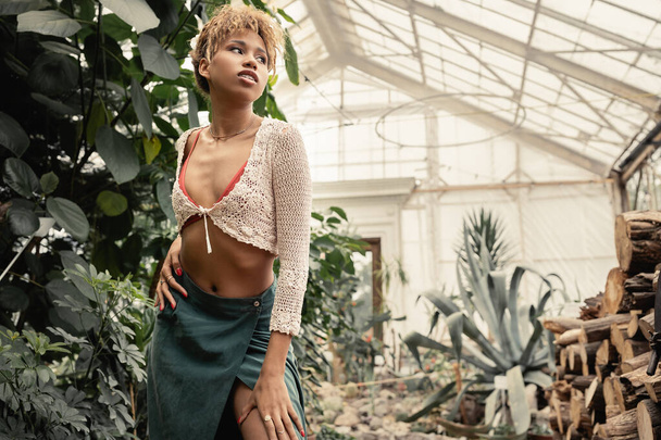 Fashionable young african american woman in summer outfit touching hip and looking away while standing in blurred garden center, stylish woman with tropical backdrop - Fotografie, Obrázek