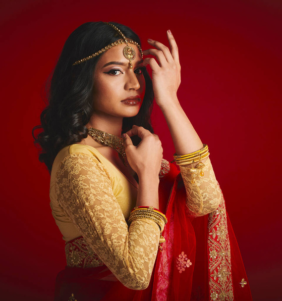 Portrait, celebration and Indian woman with fashion, traditional dress and jewellery against a red studio background. Face, female person or model with cultural clothes, religion and beauty with gold. - Фото, зображення
