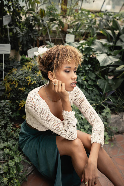 Retrato de jovem e elegante mulher afro-americana em roupa de verão relaxante perto de plantas verdes no fundo em estufa turva, senhora moda-forward em meio a vegetação tropical - Foto, Imagem