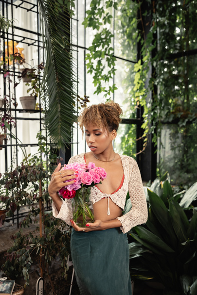 Μοντέρνο νεαρή Αφροαμερικανή γυναίκα σε πλεκτό κορυφαίο βάζο με τριαντάφυλλα και στέκεται κοντά σε θολή φυτά στο κέντρο του κήπου στο παρασκήνιο, μοντέρνα γυναίκα με τροπικό ταλέντο, έννοια του καλοκαιριού - Φωτογραφία, εικόνα