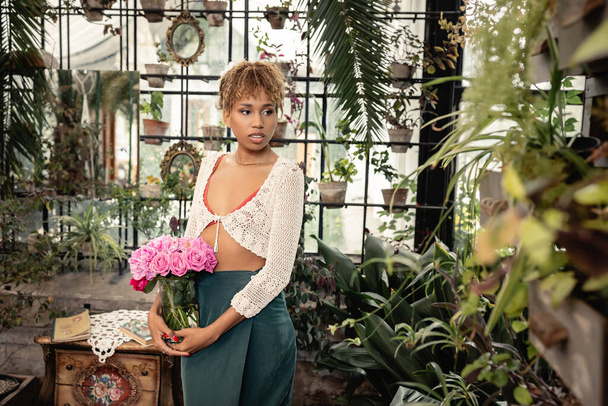 Fiatal elegáns afro-amerikai nő nyári ruhában gazdaság váza rózsaszín virágok közelében álló zöld növények a modern narancs a háttérben, trendi nő trópusi hangulat, nyári koncepció - Fotó, kép