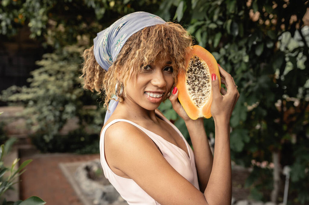 Tyytyväinen nuori afrikkalainen amerikkalainen nainen hammasraudat yllään kesämekko ja huivi tilalla tuoretta papaija ja katsot kameraa orangery, tyylikäs nainen sekoittamalla muoti ja luonto, kesäkonsepti - Valokuva, kuva