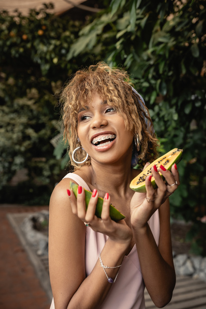 Positive young african american woman with braces in summer dress holding fresh papaya and looking away while standing near blurred plants in orangery, inspired by tropical plants, summer concept - Photo, Image