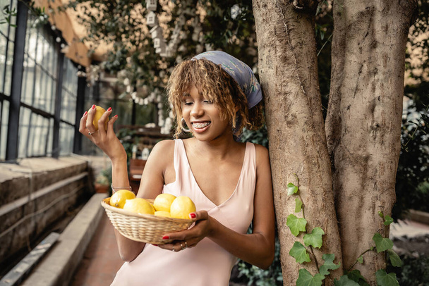 Uśmiechnięta młoda Afroamerykanka z aparatem ortodontycznym ubrana w letni strój i trzymająca koszyk z soczystymi cytrynami stojąca obok drzew w oranżerii, modna pani w harmonii z tropikalną florą - Zdjęcie, obraz