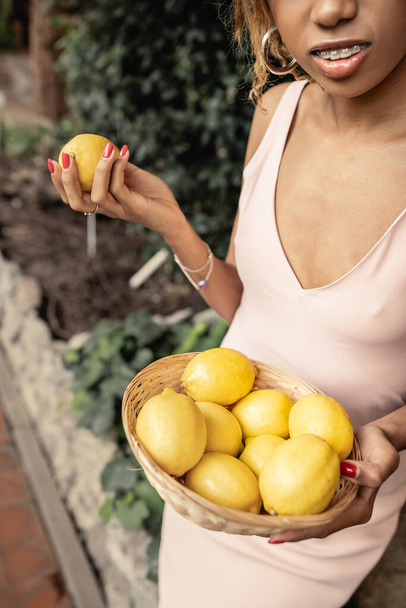 Oříznutý pohled na mladou africkou Američanku s rovnátky v letních šatech a držení šťavnatých citronů v košíku v zahradním centru, módní dáma v harmonii s tropickou flórou - Fotografie, Obrázek