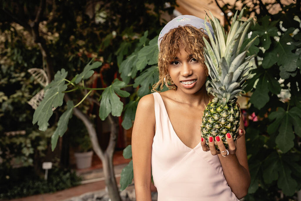 Cheerful young african american woman with braces in summer outfit holding juicy pineapple and looking at camera in blurred orangery, woman in summer outfit posing near lush tropical plants - Photo, Image
