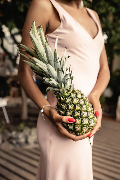 Vue recadrée d'une jeune femme afro-américaine floue en robe d'été tenant de l'ananas frais et debout dans une orangerie floue, femme élégante portant une tenue d'été entourée de feuillage tropical - Photo, image