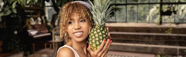 Glimlachende jonge Afro-Amerikaanse vrouw met beugel in hoofddoek en ananas vasthoudend en staand in een binnentuin, stijlvolle vrouw in zomerkleding omringd door tropisch gebladerte, spandoek  - Foto, afbeelding