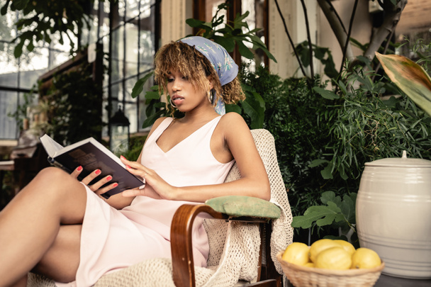 Trendy young african american woman in summer outfit reading book while sitting near blurred lemons in indoor garden, woman wearing summer outfit surrounded by tropical foliage, summer concept  - Foto, Imagen