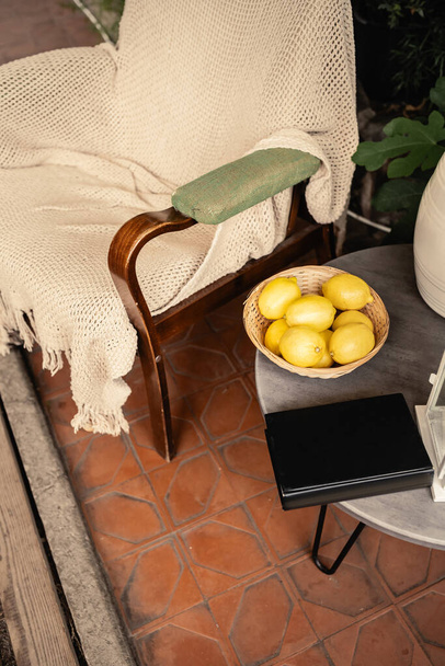 Vista de alto ángulo del libro y limones frescos en un tazón cerca de un sillón y plantas borrosas en naranjería, vitamina c, comida agria, frutas dentro del jardín interior, concepto de verano - Foto, imagen
